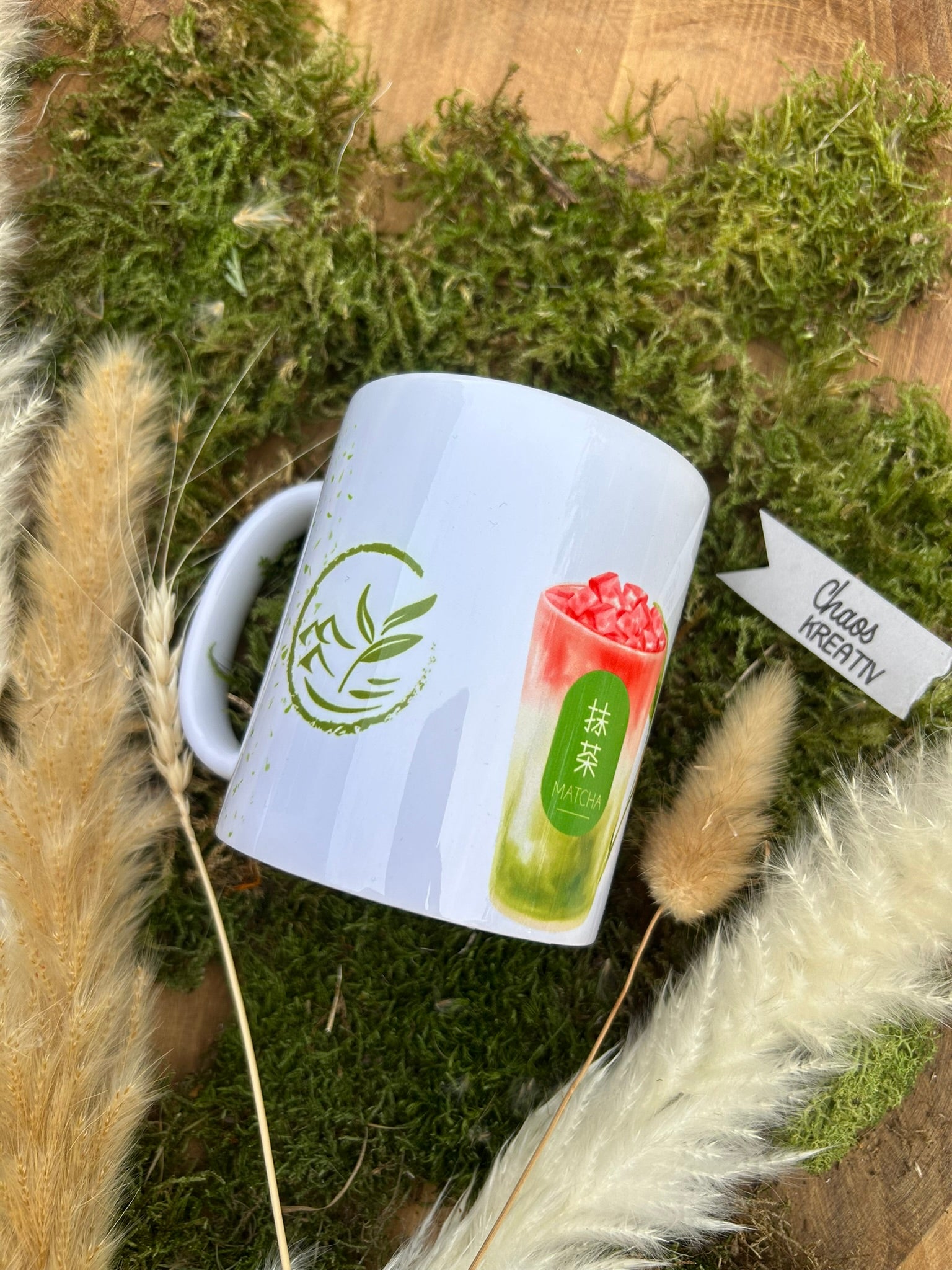 Personalisierbare 330ml Matcha Motiv-Tasse seitlich auf rustikalem Holz, umrahmt von Pampasgras und Moosdeko
